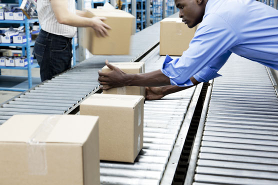 Small corrugated shipping boxes are moving down a conveyor system, with people on either side placing the boxes on it.
