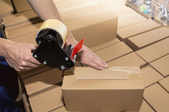 A tape gun is being used to apply clear packing tape to a small cardboard box to seal it for shipping.